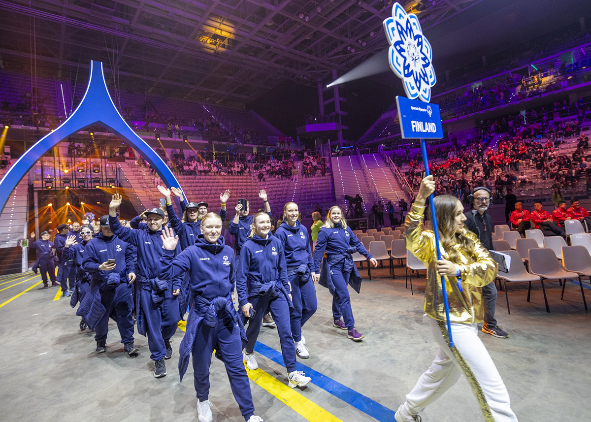 Suomen joukkue Sisujengi saapumassa areenalle avajaiskulkueessa. Edellä kävelee kultaiseen asuun pukeutunut toimitsija kantaen kisalogon lumihiutaleen muotoista Finland-kylttiä.