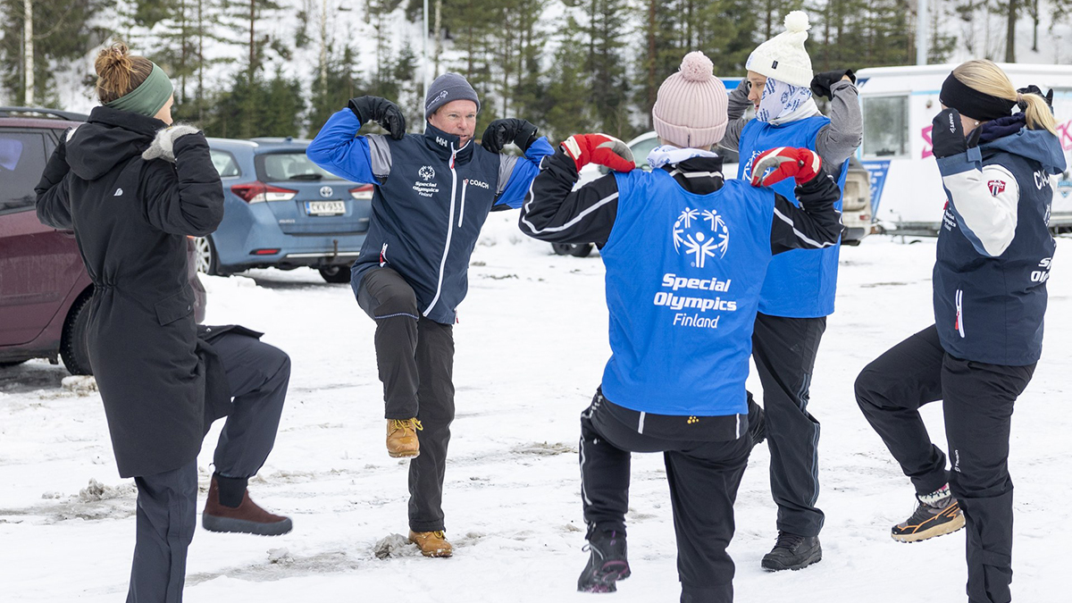 Lumikenkäilyvalmentaja Mika Valavuori vetämässä lämmittelyä lumisella kentällä. Urheilijat Janette Teerenmaa ja Aleksi Timonen sekä leirin vetäjiä nostelemassa käsiä ja jalkoja mallin mukaan.